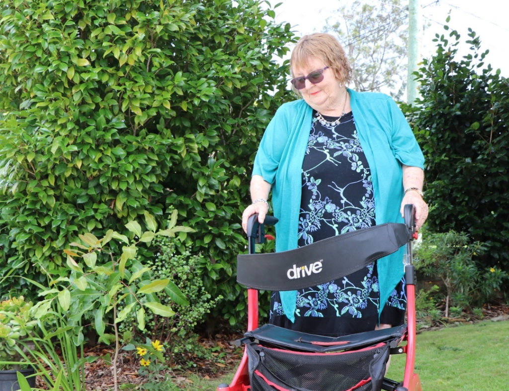 Link Wentworth resident Anne wheels her red and black walker on the grass and looks down at her garden. She is wearing sunglasses, a blue cardigan and a patterned navy shirt.