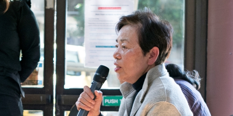 Elderly woman with short hair speaking into a microphone while sitting down at an event. She is wearing a grey jumper with a high collar.